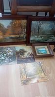 All six artworks displayed together: two large framed paintings leaning, three unframed prints and one small framed seascape on floor.