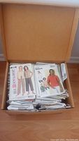 Full open cardboard box filled with folded paper sewing patterns showing a variety of designs including pants, tops, and jackets.