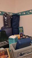 Photo showing camping items including sleeping bags, cooler, boxed bug zapper, propane bottles, stove and solar shower packaging.
