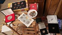Photo showing multiple coin sets on wooden surface including boxed specimen set, coins in plastic, red velvet display case, and loose coins in plate.