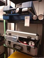 Photo showing two closed forensic equipment cases stacked, one blue plastic and one silver metal, with flashlight and measuring wheel handles visible.