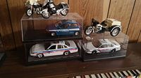 Wide view showing three police die-cast model cars in plastic display cases and three police motorcycles on display stands arranged on wooden surface against wood panel background.