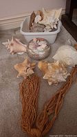 Photo shows large seashells, a piece of driftwood, and a pair of brown macrame hangers on the floor next to the plastic bin.