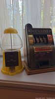 Overall photo of yellow plastic gumball machine with clear globe and Nevada slot machine with wood base beside it