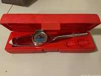 Open red molded plastic case containing chrome Snap-On torque meter with dial and red handle, missing sockets.