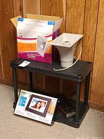 View of Radio Shack Powerhorn speaker with its box, the Coby digital photo frame box visible, and the plastic business card organizer shelf unit on carpet against wood panel wall.