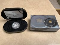 Black oval metal presentation case open showing the silver coin with the Bluenose ship and certificate inside, next to the outer box.