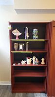 Full front view of the wooden five shelf bookcase with some glass and ceramic decorative items placed on the shelves, showing scratches on the wood.