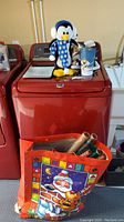 Penguin plush on top of red washing machine with bag of Christmas wrapping paper beneath