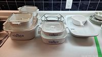 Nine assorted Corning Blue Cornflower dishes and plates with lids on a kitchen counter.