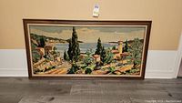 Full view of the framed crewel embroidery artwork showing a landscape scene with buildings, trees, and water.