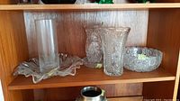 Six different crystal and glass vessels displayed on wooden shelf including vases and bowls showing shape, size, and pattern.