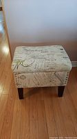 Front and top view of a rectangular foot stool upholstered in cream fabric with vintage script print, showing cushion top and wooden legs.