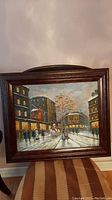 Full view of the framed canvas painting showing a snowy city scene with people and buildings, displayed on a striped cloth surface.