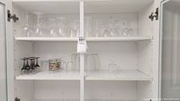 Wide view of white cabinet shelves holding assorted clean glassware including upside down wine glasses, pint glasses, shot glasses, and small jars.
