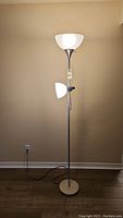 Full view of the two-bulb standing lamp, front facing with both bulbs visible. Lamp is plugged in and standing on a hardwood floor against a beige wall.