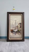 Front view of original acrylic framed painting depicting boats docked at a pier by buildings with cloudy sky.