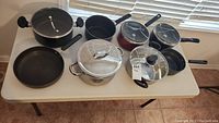 Eight cookware pieces including three black pots with glass lids, one large stainless steel pot with glass lid, and multiple black frying pans without lids placed on tan table by window.