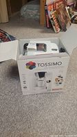 White Tassimo T55 single cup coffee brewing system in open box on carpeted floor showing front and top views