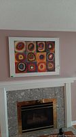Front view showing entire framed abstract artwork with 12 colorful concentric circle panels hung above a fireplace.