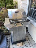 Full side view of gas grill showing stainless steel hood, two control knobs, handle, and base with door and vent.