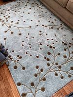 Photo showing overall rug laid on wood floor near sofa, highlighting aqua color and scattered brown botanical elements