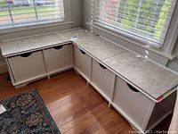 Two cream-colored corner cubby shelving units arranged in an L-shape in front of windows, showing their marble-look top surfaces and two drawer fronts each with cutout handles.