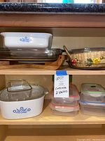 Overview of two wooden kitchen shelves with Corning Ware casseroles, glass bakeware, Longaberger basket with flowers, and Sterilite food storage containers