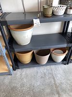 Black plastic shelving unit with three shelves holding flower pots (not included in sale). Shows shelves and tubular leg construction.