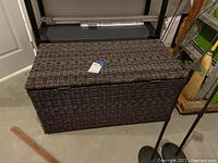 Photo showing large brownish/grey wicker-style PVC storage chest from a frontal corner angle, displaying the woven pattern and handles on the side.