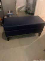 Closed navy storage ottoman with studded trim on the side and four black tapered legs.