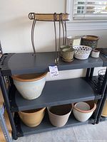 Photo showing a shelving unit with multiple ceramic and plastic flowerpots of various sizes and two metal plant stands on the top shelf.