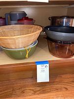 Wide shot showing green and clear Pyrex mixing bowls stacked, wooden woven basket on top, Circulon frying pan on top of a brown glass Pyrex bowl, and stainless steel stockpot in the background.