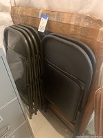 Photo showing six folded black folding chairs with padded seats and backs, stacked together beside a cardboard-wrapped item presumed to be the round folding table.