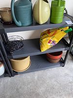 Wide view of plastic shelving unit with three shelves. Various garden and household items present but not included in sale.