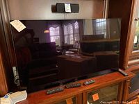 Frontal view of glossy black bezel Sony TV on wooden console with three remotes and one headset visible.