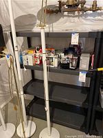 Full view of the heavy duty utility shelf with four shelves, placed against a basement wall underneath pipes. Shelf holds various small items in baskets and boxes. Dark gray metal construction visible. Shelf dimensions and tags visible.
