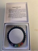 Top view of the onyx chakra bracelet inside a white box, showing black beads and seven colorful chakra stones, with an informational card on chakra properties.