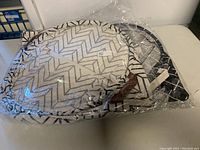 Three brand new folded canvas collapsible baskets, still inside clear plastic packaging, stacked on a white surface. Different geometric patterns visible on each basket.