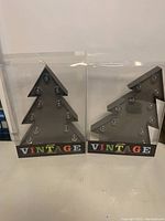 Two metal vintage marquee Christmas trees side by side, showing front view with clear light bulbs and vintage marque text on packaging.