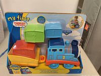 Photo of packaged toy set showing blue Thomas engine, orange train car, and three colorful stackable blocks in green, blue, and orange.