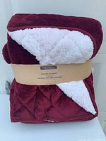 Folded quilted blanket in burgundy with visible white Sherpa backing showing, label attached.