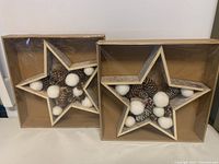 Two boxed rustic farmhouse style star decorations with pine cones and white pom-poms inside, visible through packaging.