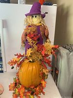 Full view of fabric scarecrow standing on top of a pumpkin decorated with faux autumn leaves.