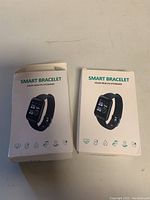 Photo of two boxed smart bracelets, one box slightly squished, showing front image of bracelet with black band and screen displaying time and icons.