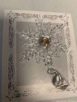 Close up on a clear snowflake with an amber gem in center and dangling clear gem, mounted on decorative backing