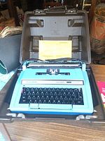 Blue Smith Corona Electra C-T electric typewriter inside an open brown hard plastic case with a yellow instruction manual visible behind the typewriter.