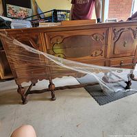 Full side view of antique solid wood sideboard wrapped in plastic, showing ornate carved front panels on drawers and doors and turned legs.