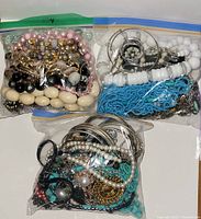 Photo showing three clear plastic bags filled with assorted costume jewelry including necklaces, bracelets, earrings, and bangles in various colors and materials.