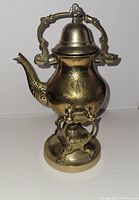 Front view of ornate brass Middle Eastern teapot with handle, spout, lid, and decorative engravings including 'Saudi Arabia' inscription. Shows the wick burner base.